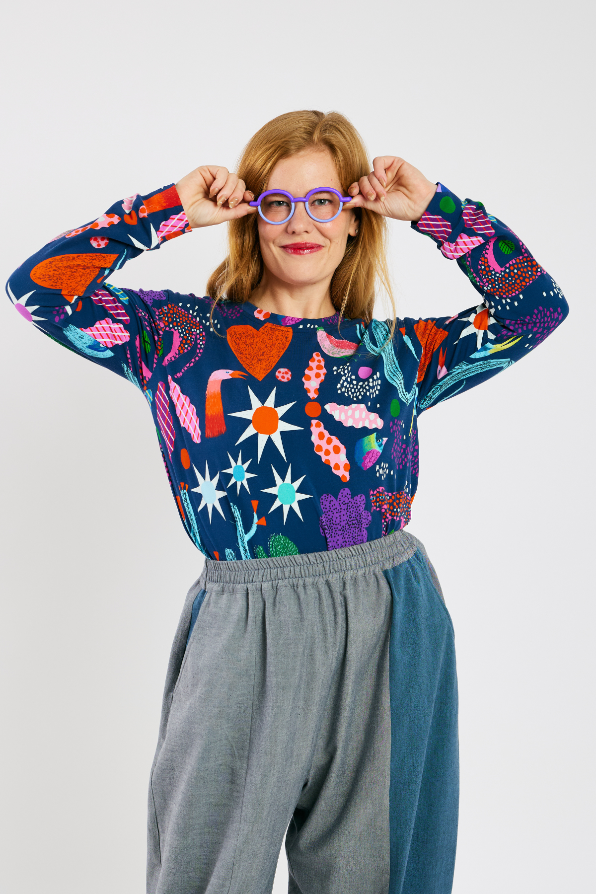 Woman wearing a colorful patterned shirt with a bright pattern, glasses from Eye Spy Optical,  and sustainable gray pants on a white background