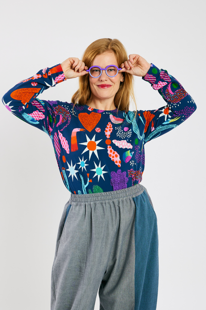 Woman wearing a colorful patterned shirt with a bright pattern, glasses from Eye Spy Optical,  and sustainable gray pants on a white background