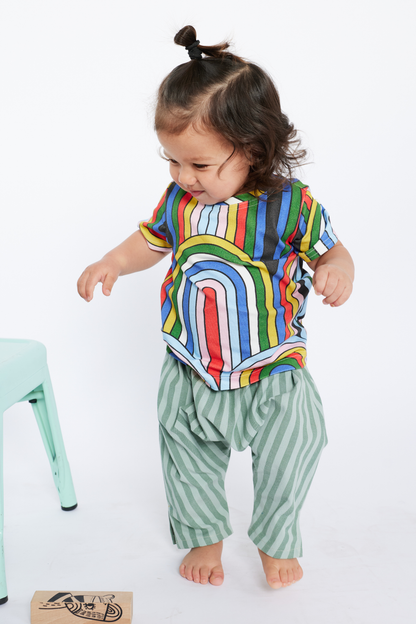 Child wearing a colorful rainbow shirt and green striped pants by Lilla Barn Clothing on a white background