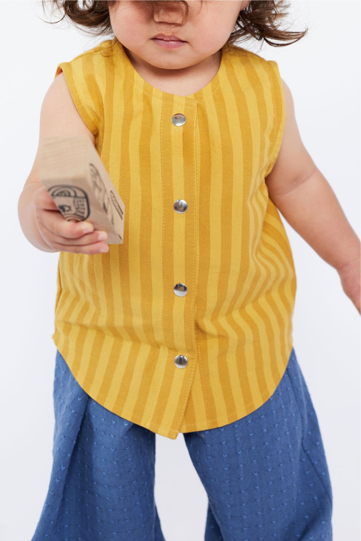 Child wearing a yellow striped shirt and blue pants holding a wooden block.