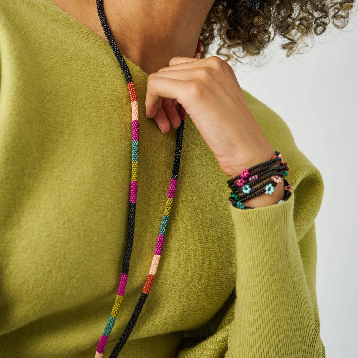A close-up lifestyle shot of a person wearing a chunky black beaded necklace with colorful geometric bead segments (pink, orange, yellow, green, blue) and matching beaded bracelets on the wrist, styled with an olive green knit sweater.