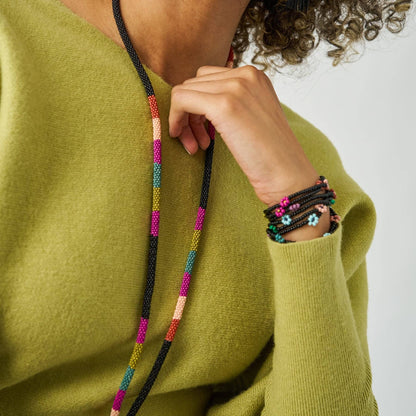 A close-up lifestyle shot of a person wearing a chunky black beaded necklace with colorful geometric bead segments (pink, orange, yellow, green, blue) and matching beaded bracelets on the wrist, styled with an olive green knit sweater.