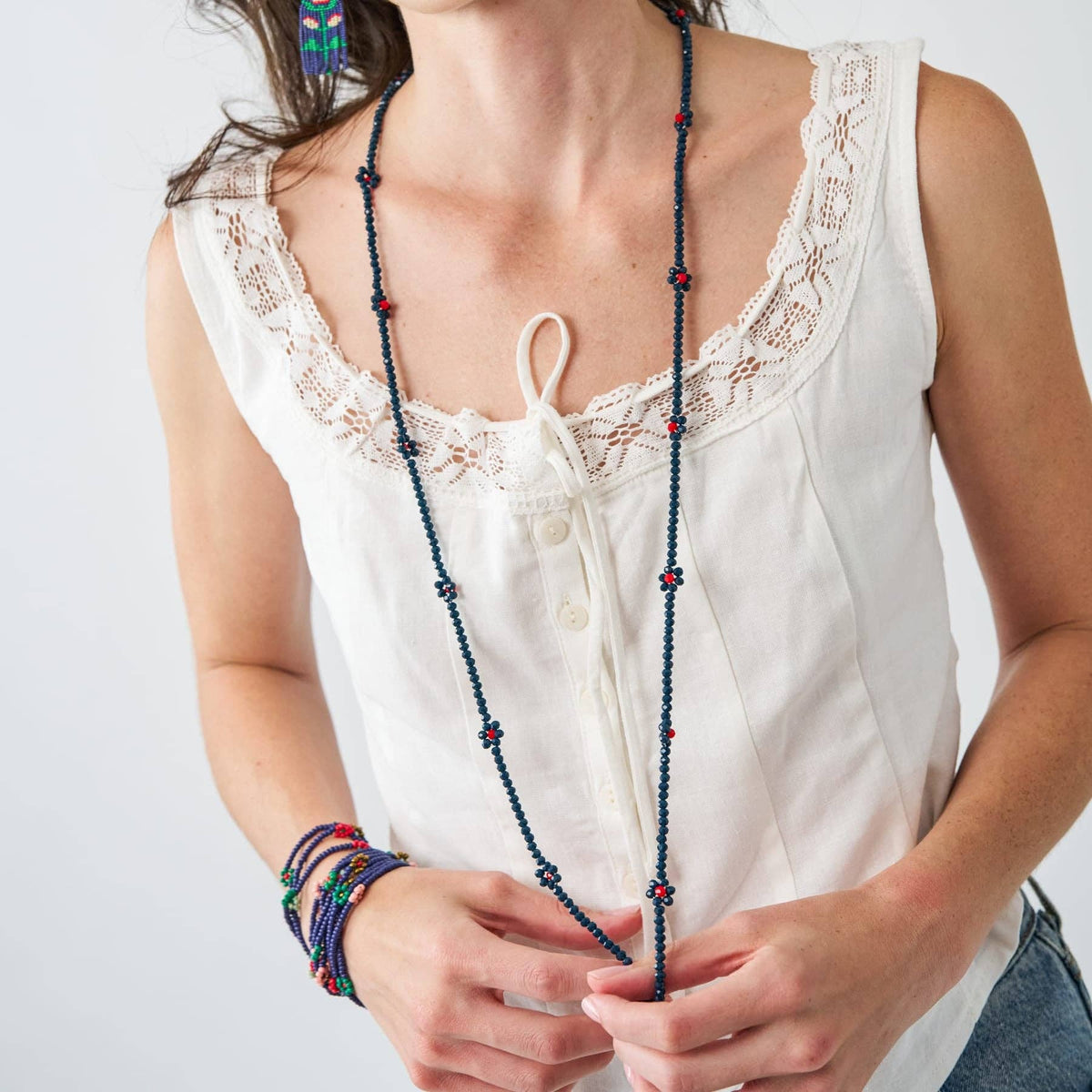 A woman wearing a long beaded necklace with dark blue beads and small red accents, styled with a white lace-trimmed top and colorful bracelets.