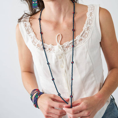 A woman wearing a long beaded necklace with dark blue beads and small red accents, styled with a white lace-trimmed top and colorful bracelets.