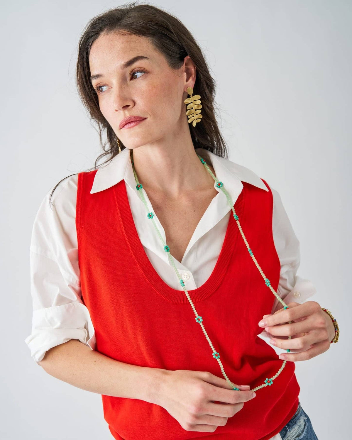 A woman wearing a bright red knit vest over a white collared shirt, accessorized with a long beaded necklace and gold statement earrings, photographed against a light gray studio background.