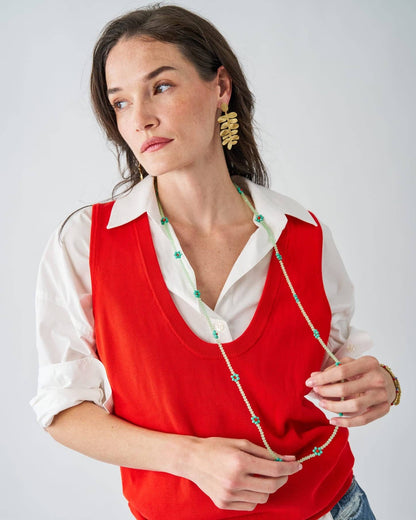 A woman wearing a bright red knit vest over a white collared shirt, accessorized with a long beaded necklace and gold statement earrings, photographed against a light gray studio background.