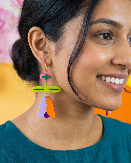 A close-up of a woman wearing colorful geometric dangle earrings with abstract flower-like shapes in green, orange, purple, and pink.