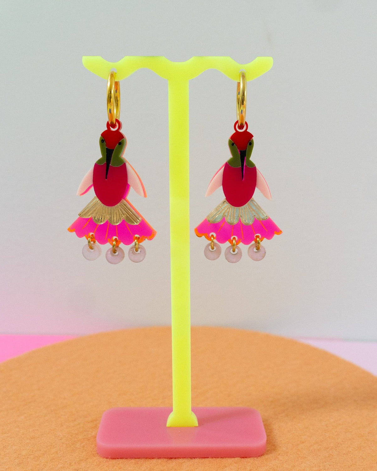 A pair of colorful dangling earrings displayed on a bright yellow stand. Each earring features a red top section with green and pink details, a pink patterned skirt, and small clear bead drops.