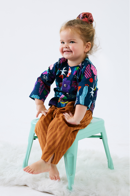 Child sitting on a small stool wearing a colorful shirt and brown stripe pants against a white background