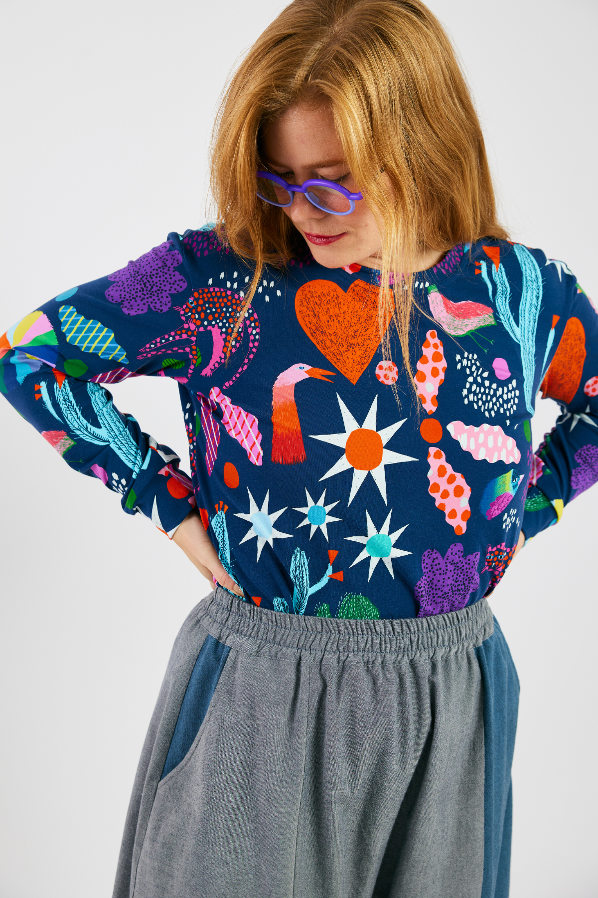 Woman wearing a colorful patterned shirt with a bright pattern, glasses from Eye Spy Optical,  and sustainable gray pants on a white background