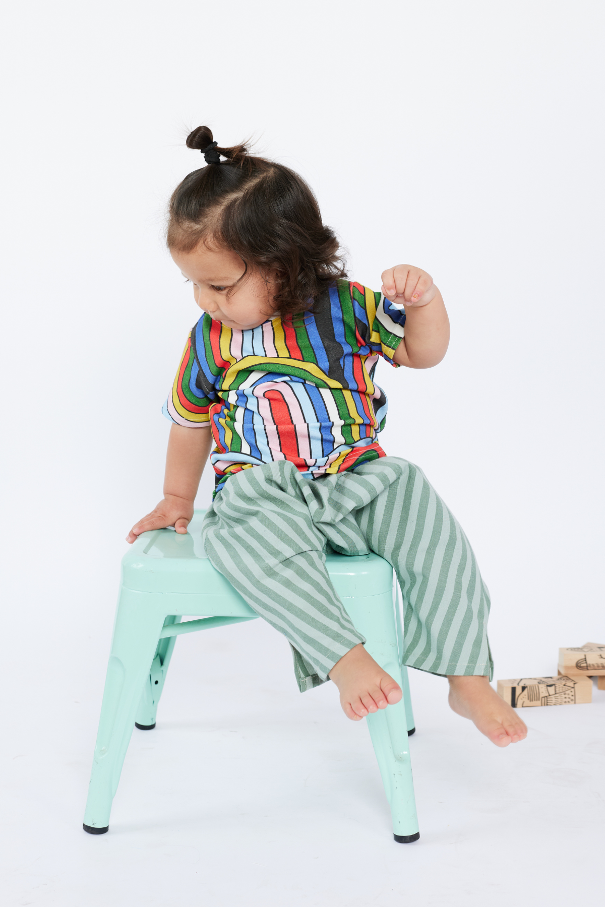 Child wearing a colorful striped shirt and green pants sitting on a small stool against a white background