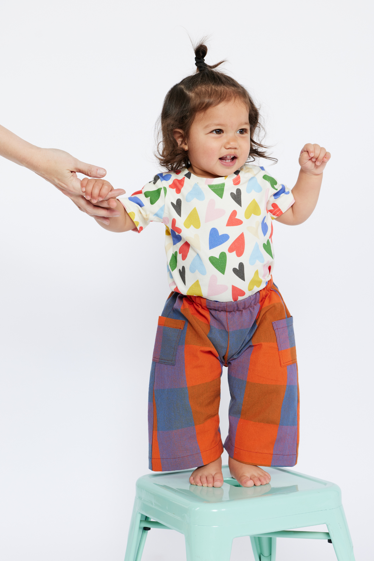 Child wearing a colorful shirt with heart patterns and checkered pants standing on a stool.