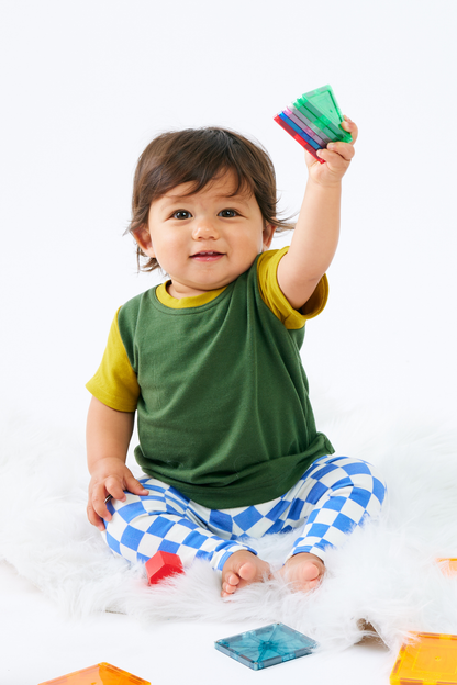Baby holding colorful magnatiles and wearing a green and yellow bamboo t-shirt with blue checkered organic cotton leggings, sitting on a white surface.