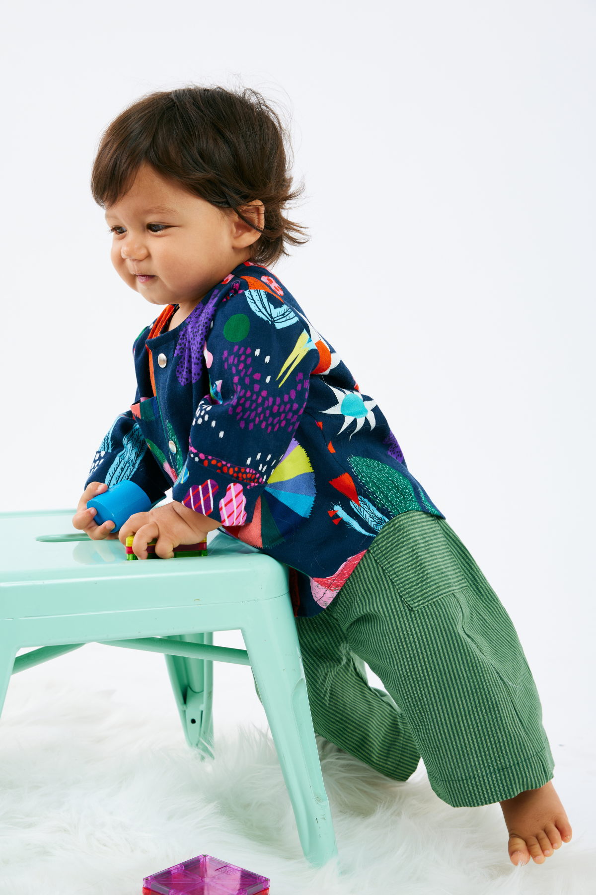 Child wearing a colorful shirt and green stripe pants with pockets - playing with a colorful toy on a white background