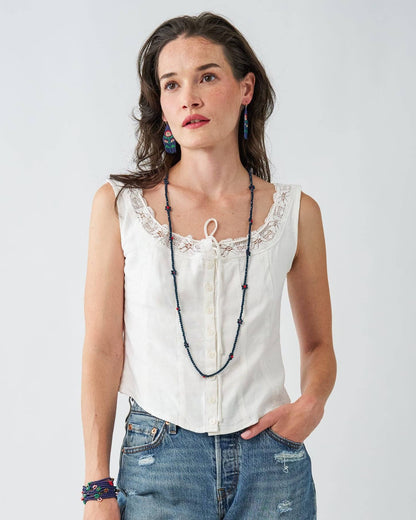 A woman wearing a sleeveless white top with lace trim and a front tie detail, paired with blue jeans and a long beaded floral necklace, against a light gray studio background.