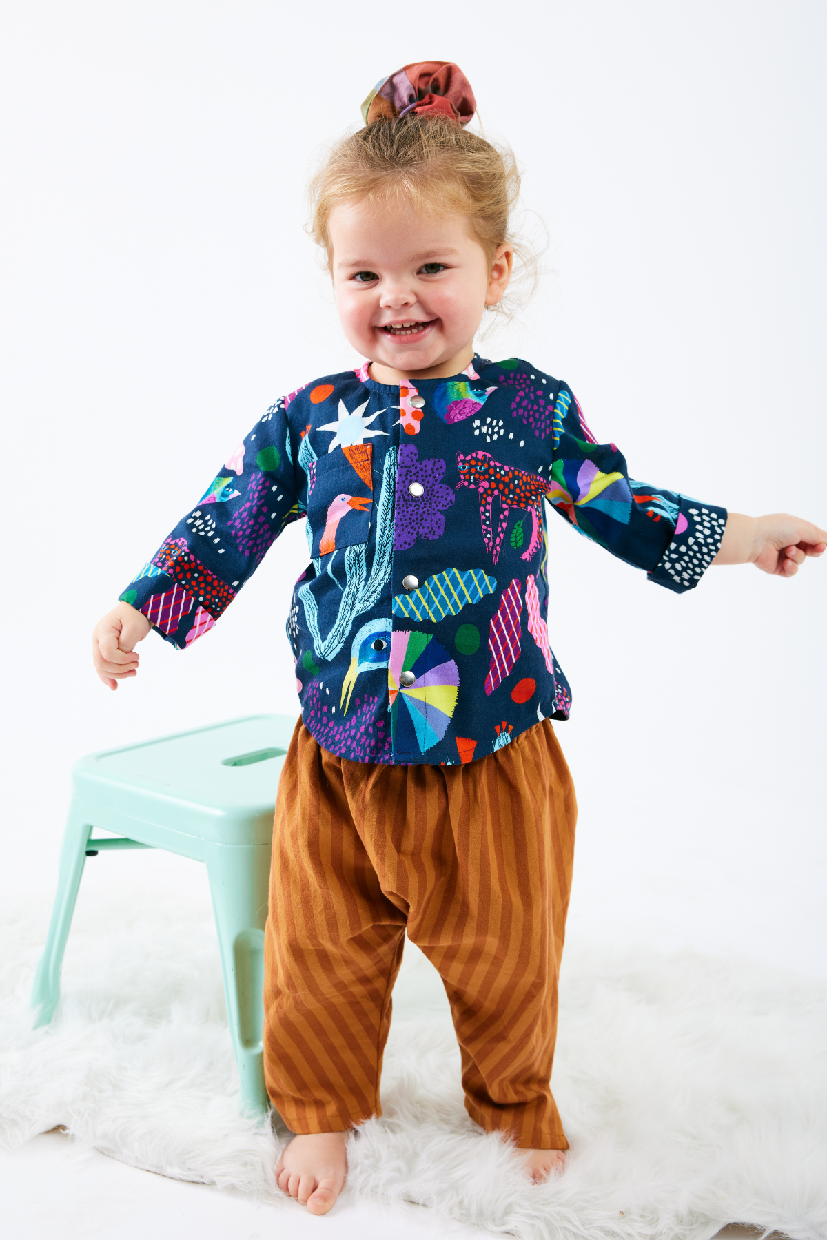 Child wearing a colorful long-sleeve shirt with abstract patterns and brown striped pants on a white background.