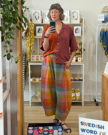 A woman takes a mirror selfie in a boutique while wearing a rust red short-sleeve button-up shirt and vibrant, multicolored plaid wide-leg pants. The boutique interior features white shelves with framed art and clothing racks.