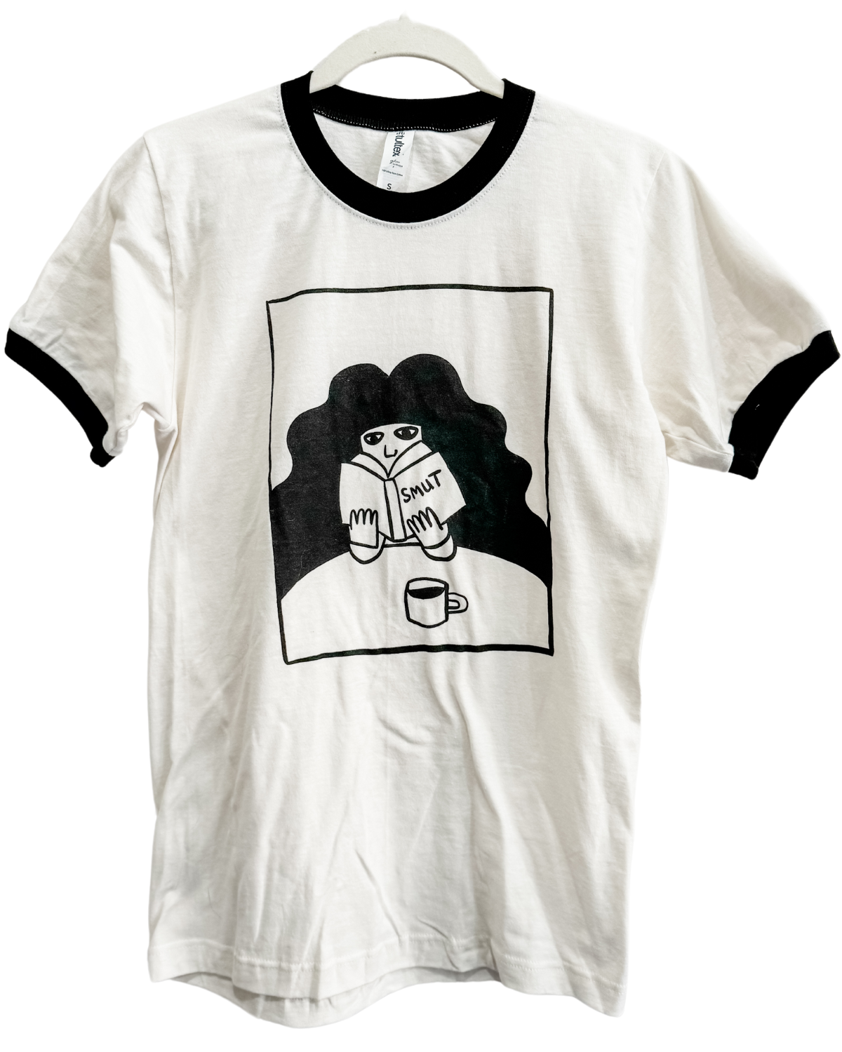 Off-white cotton tee with black neck and arm cuffs with a person reading a book labeled smut and drinking a cup of coffee 