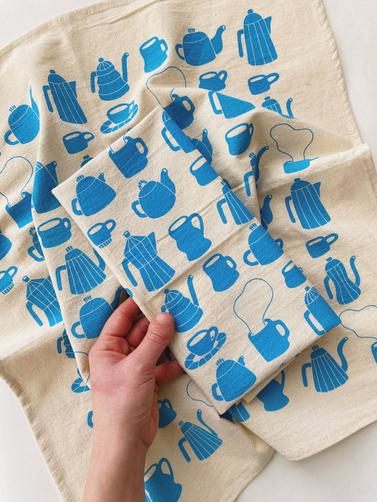 Folded cotton tea towel with blue teapot pattern held by a hand on a light background