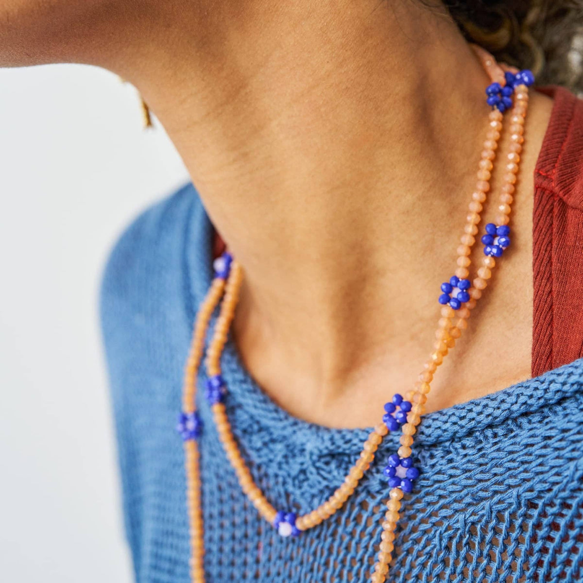A close-up of a person's neck and upper chest wearing a double-strand beaded necklace. The necklace features peach-orange beads with small blue flower-shaped accents, worn over a blue knit top and a rust-red shirt.