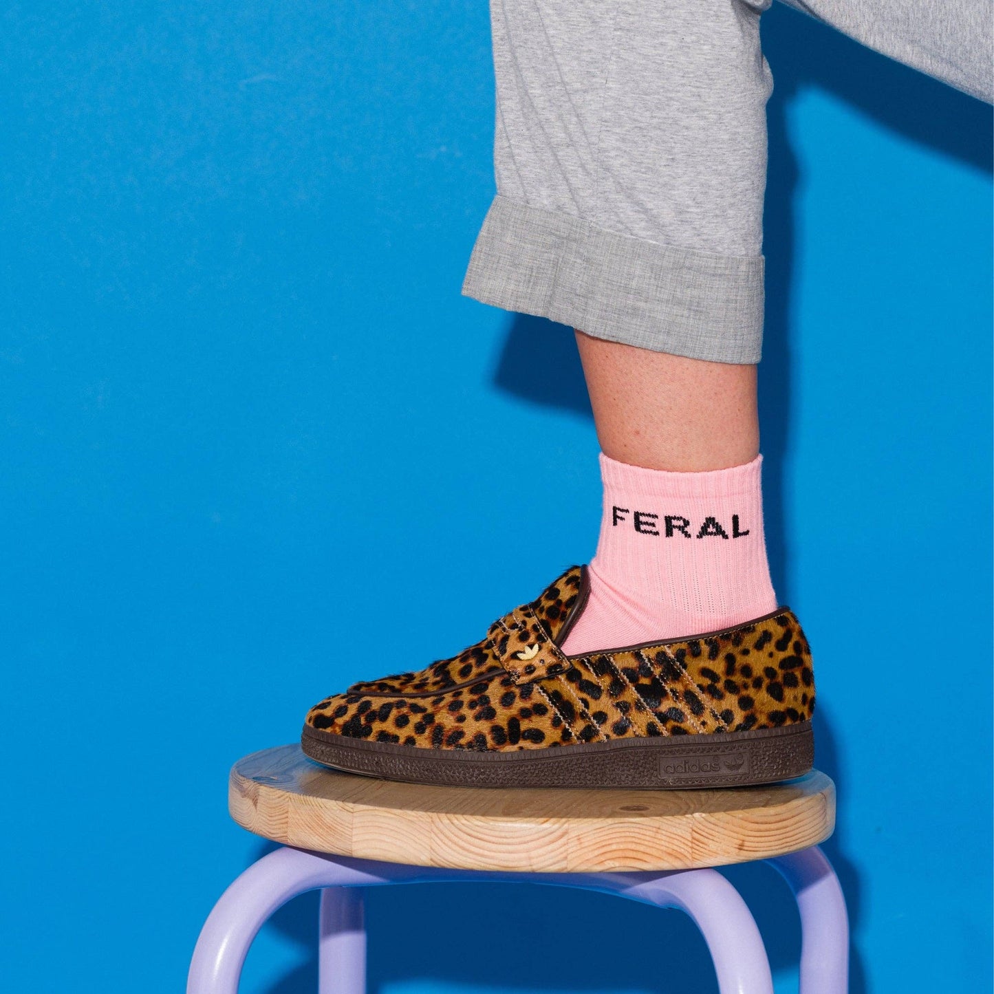 A person wearing a leopard-print slip-on shoe with a pink sock featuring the brand name 'FERAL', posed on a wooden stool against a bright blue background.