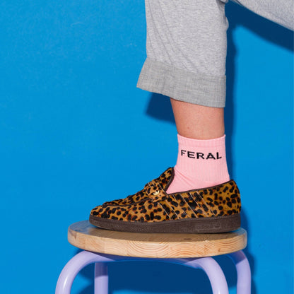 A person wearing a leopard-print slip-on shoe with a pink sock featuring the brand name 'FERAL', posed on a wooden stool against a bright blue background.
