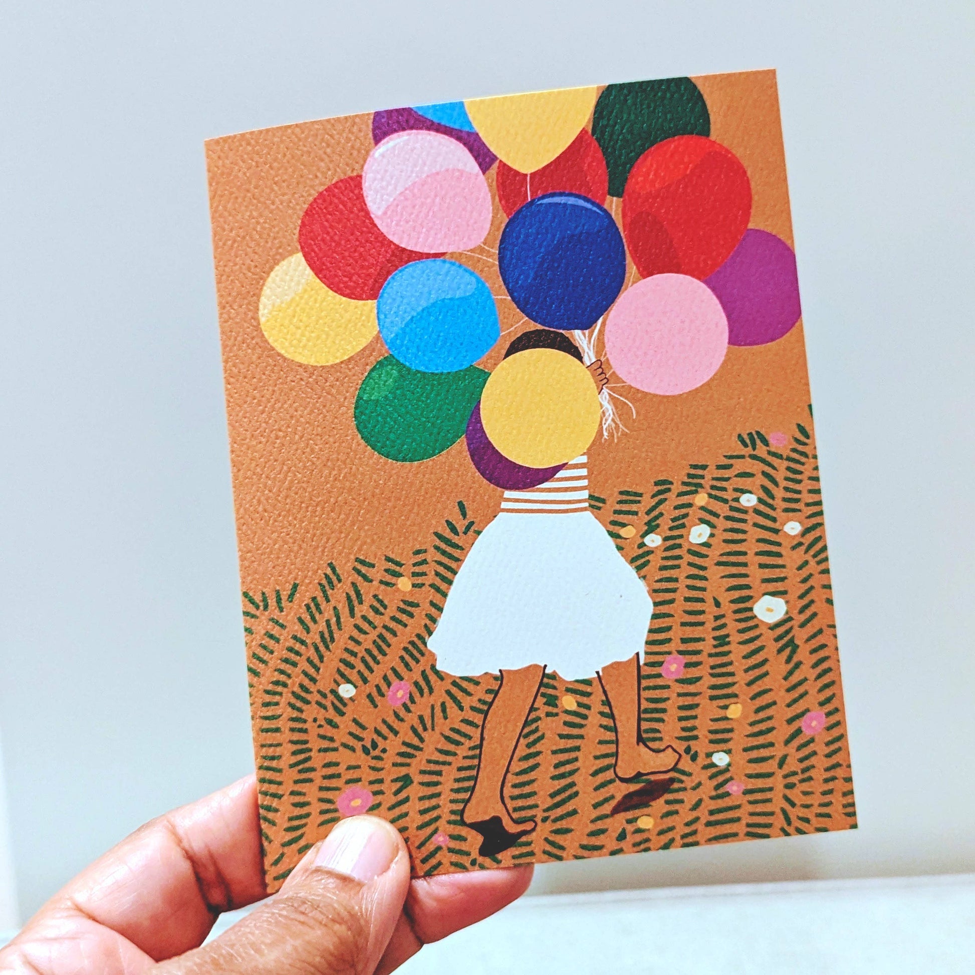 Illustrated card with colorful balloons and a person holding them, held by a hand against a light background.