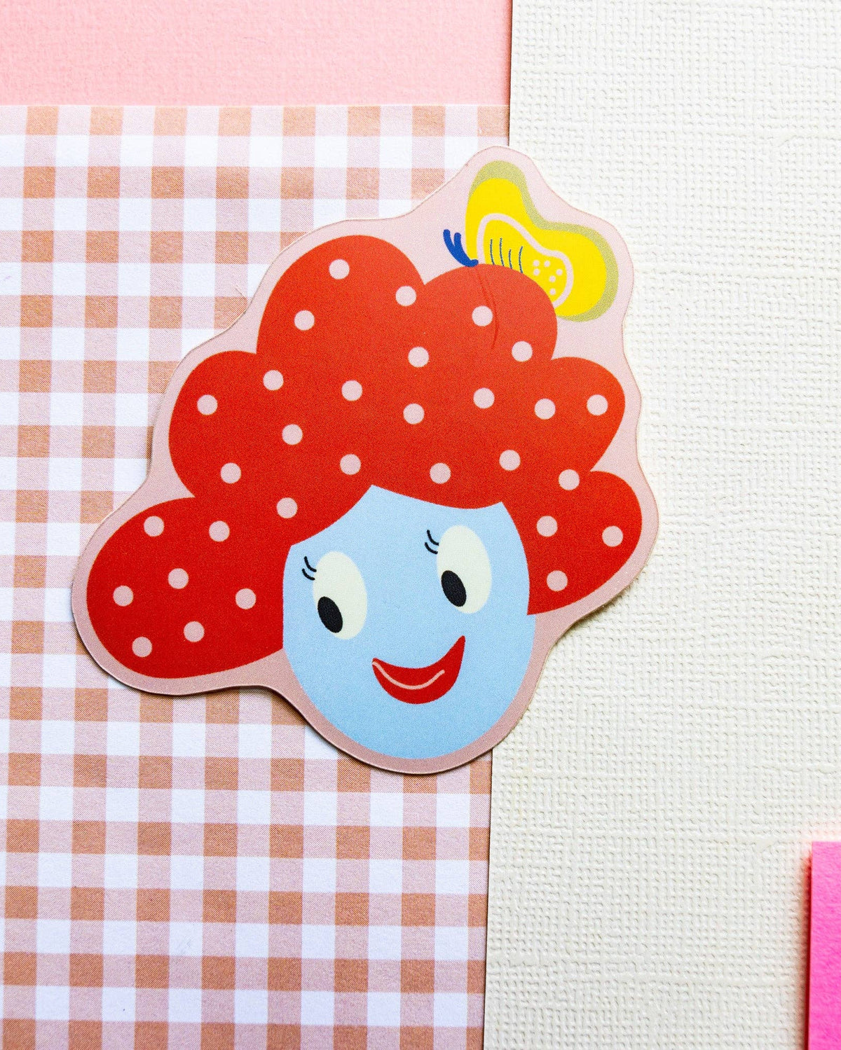 A die-cut sticker featuring a cartoon face with red polka-dot hair, a blue face, and a yellow hairbrush, shown on a pink-and-white gingham paper background.