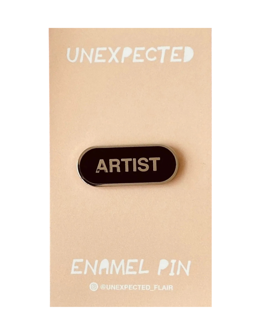Enamel pin with 'ARTIST' text on a beige card with 'UNEXPECTED' branding.
