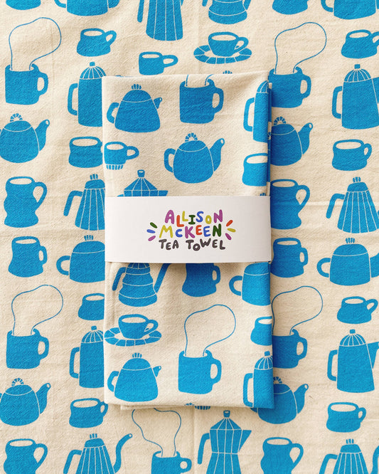 Tea towel with blue teapot and cup pattern on beige background, featuring the brand 'Allison Mckeen'.