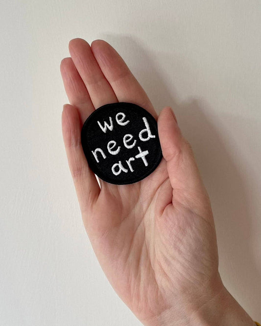Hand holding a black embroidered patch with 'we need art' text on a plain background