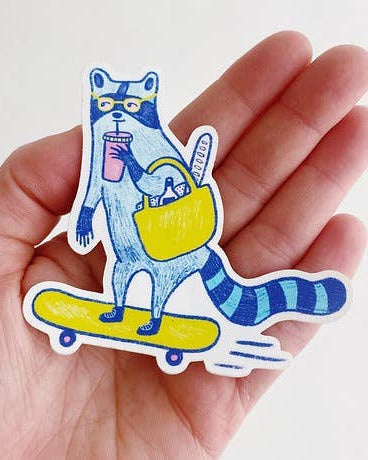 Colorful sticker of a raccoon on a skateboard held in a hand against a white background