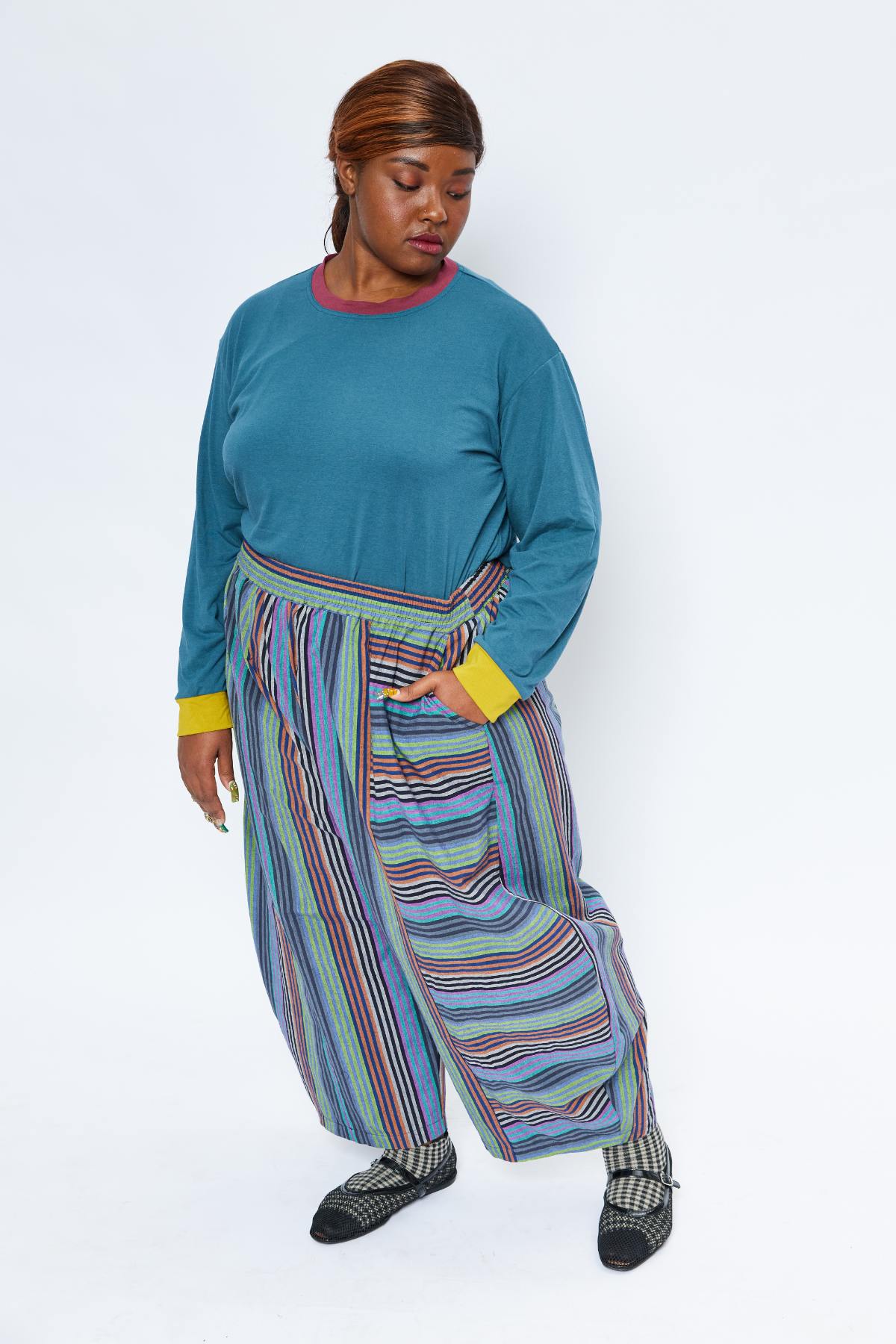 plus size woman standing with her hand in her pocket. She's wearing slow fashion garments including a blue shirt and dark striped barrel pants.