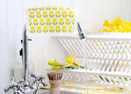 A white dish drying rack with a yellow tulip pattern, shown in a bathroom setting with running water from a chrome faucet and yellow daffodils in the background.