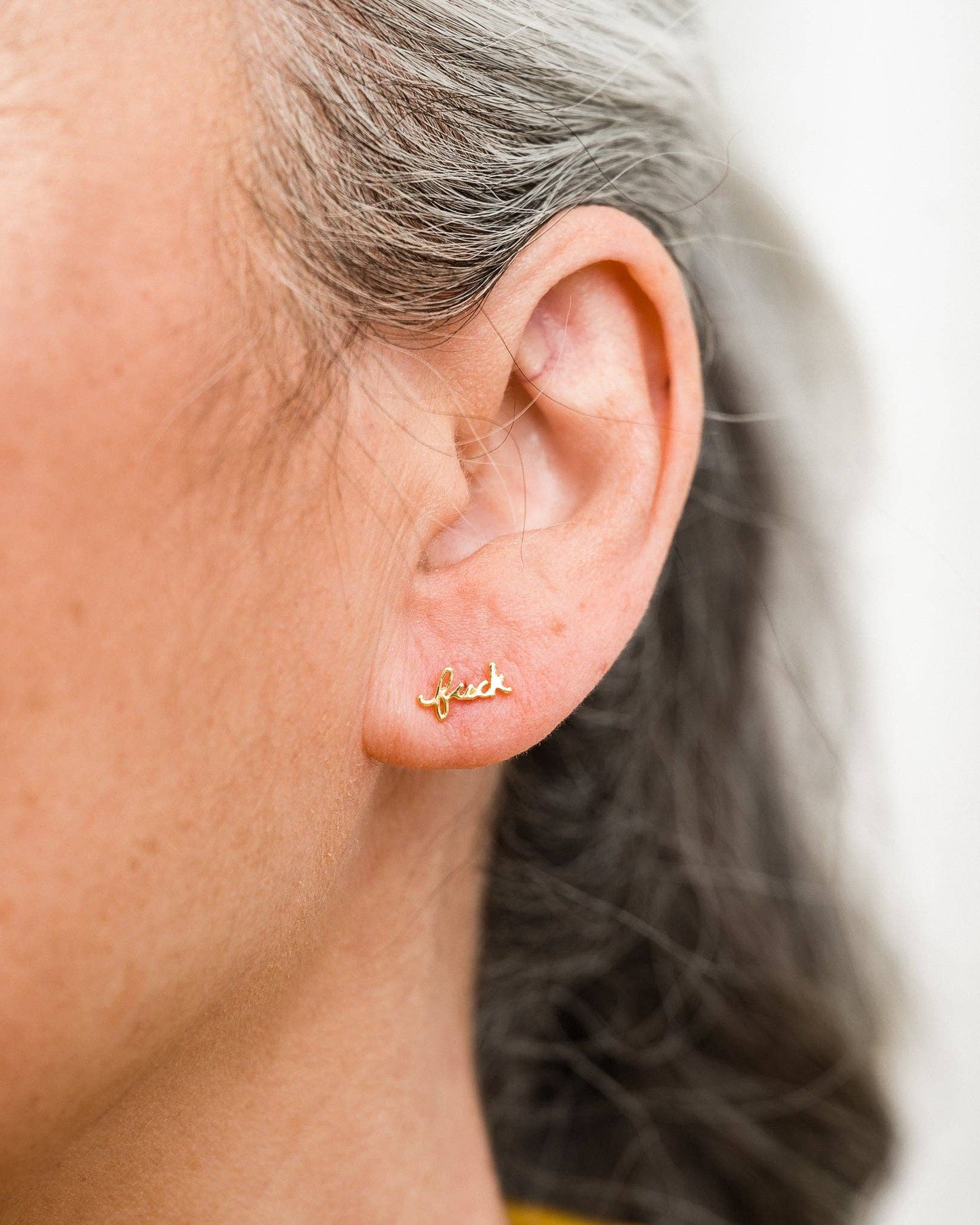 Close-up of an ear wearing a gold stud earring that says "f*ck"