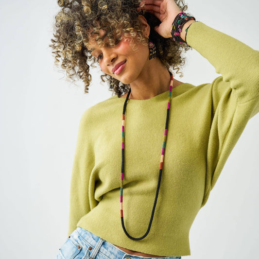 A woman wearing a bright yellow-green knit sweater and a long black beaded necklace with colorful accents, posing against a light gray background.