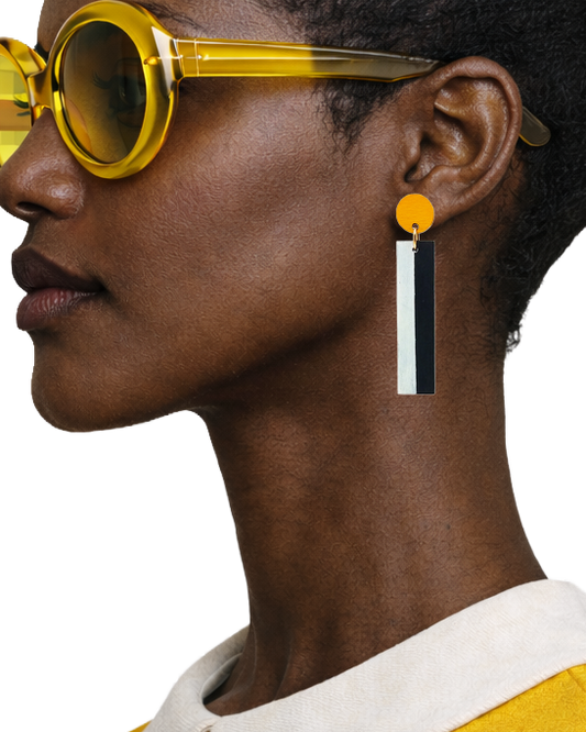 A close-up side profile of a model wearing a geometric dangle earring. The earring features a bright orange circular stud connected to a vertical bar with alternating white and black rectangular segments.