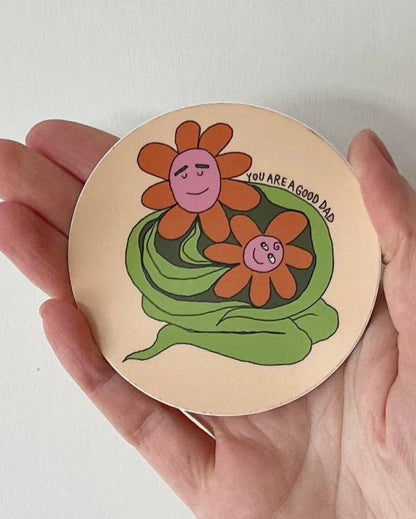 Round wooden sticker with floral design and text held in a hand against a white background