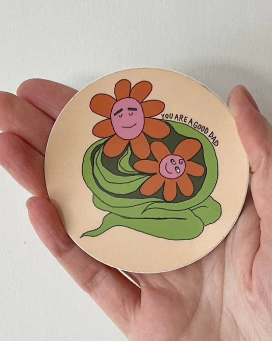 Round wooden sticker with floral design and text held in a hand against a white background