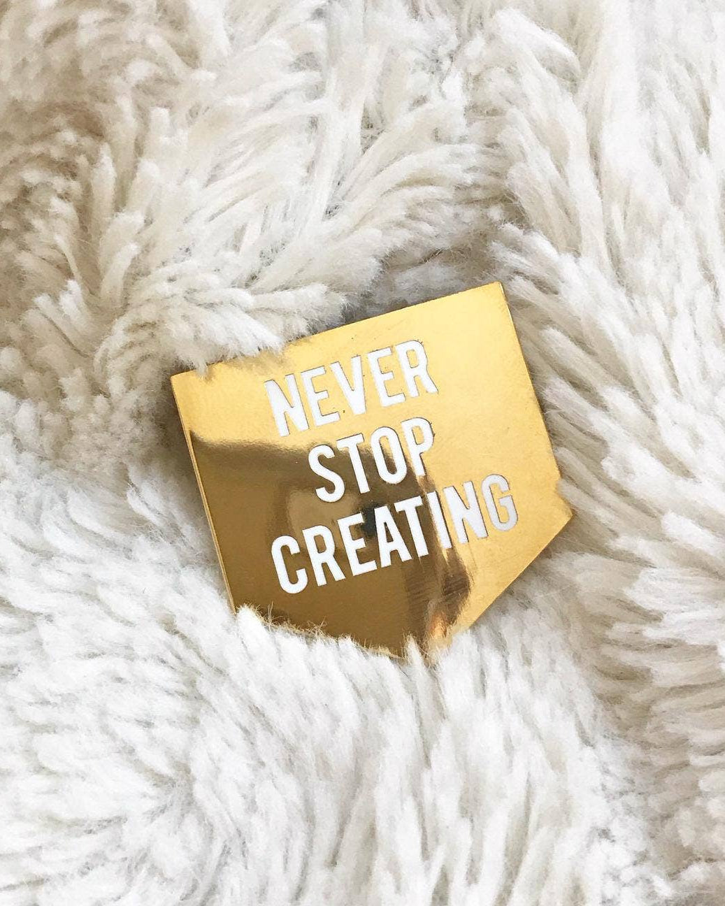 Gold enamel pin with 'NEVER STOP CREATING' text on a fluffy white surface