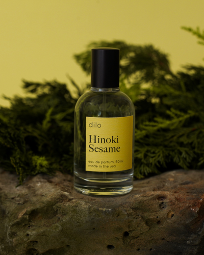 A clear glass bottle of Hinoki Sesame eau de parfum with a matte black sprayer, placed on a rock, with green pine needles in the background.