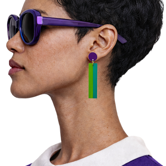 A close-up side profile of a person wearing purple sunglasses and a long geometric drop earring with a purple top circle and two vertical bars in green and blue, against a plain white background.