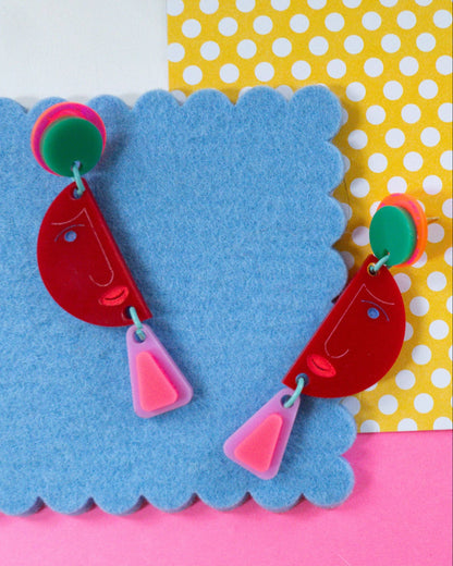 A pair of colorful geometric dangle earrings displayed on a blue scalloped felt card. The earrings feature a red crescent-shaped drop with a small blue dot, a pink triangle below, and a green and orange circular stud at the top.