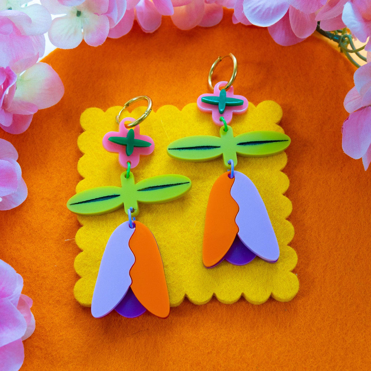 Colorful acrylic dangle earrings with a floral and leaf design, displayed on a yellow felt card against an orange background with pink flowers.