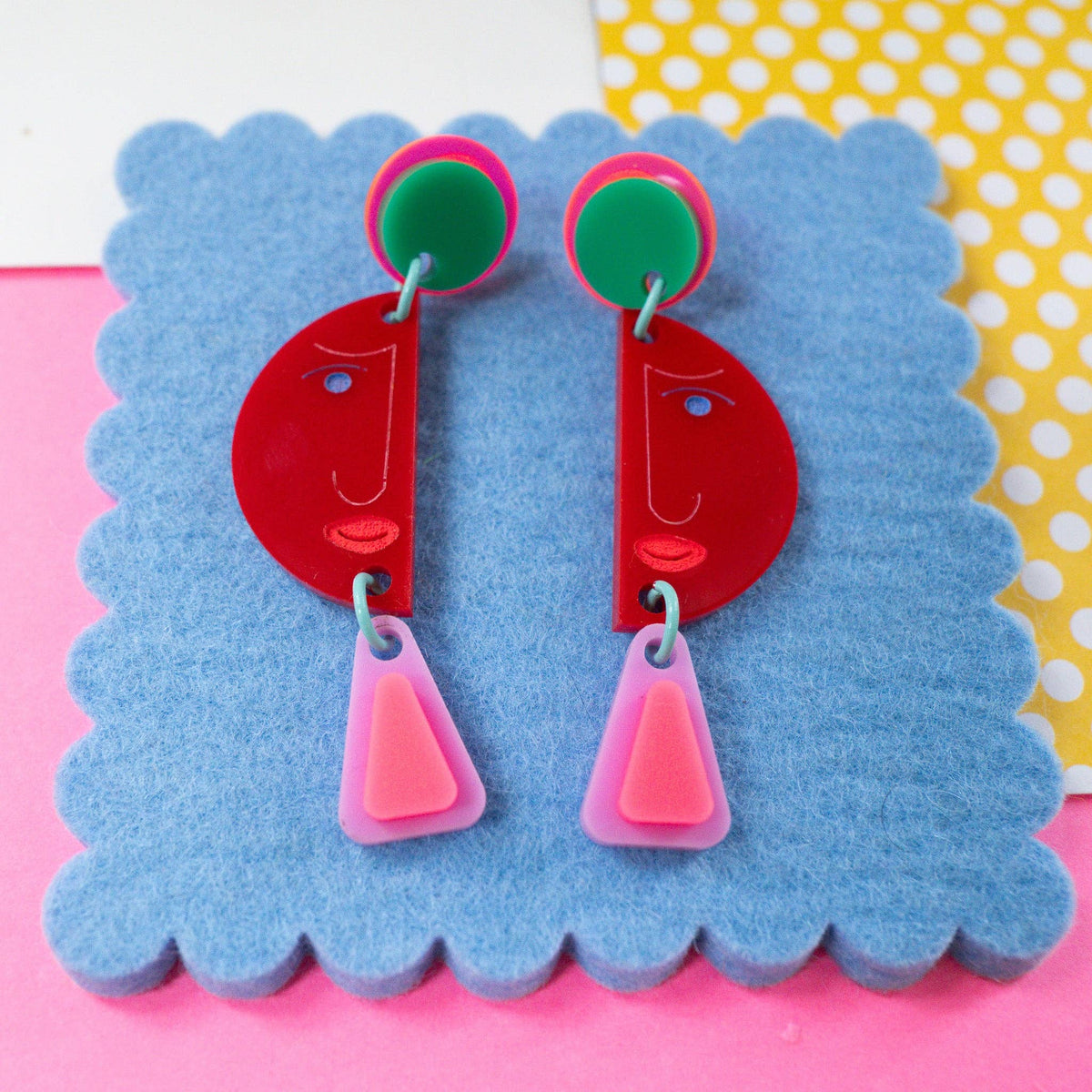 A pair of colorful acrylic dangle earrings displayed on a blue scalloped card. The earrings feature red crescent-shaped drop pendants with simple line-art facial features, hanging from bright pink and green circular studs.
