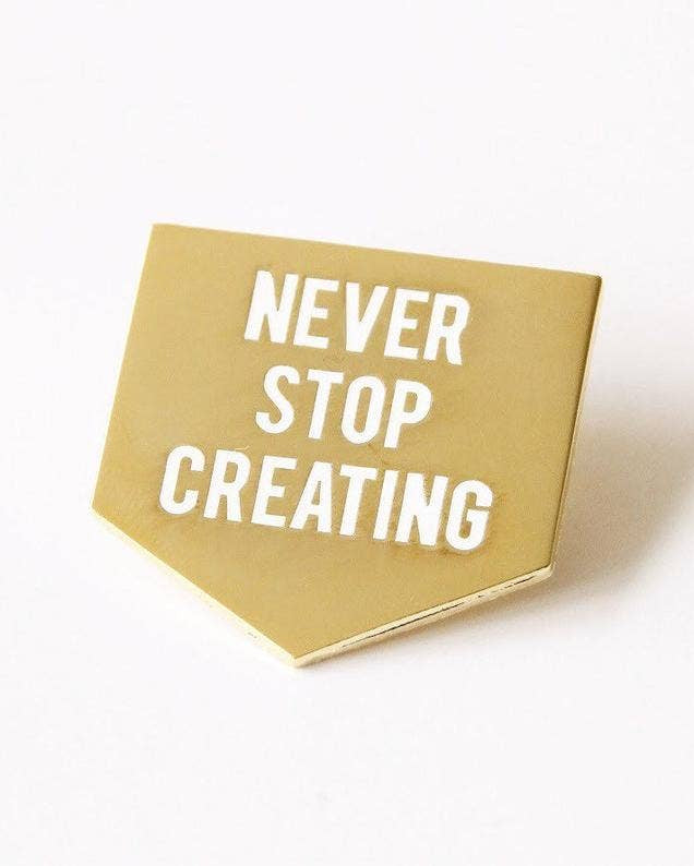 Gold enamel pin with 'NEVER STOP CREATING' text on a white background