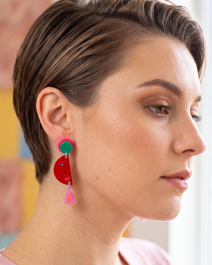 A close-up side profile of a woman with short brown hair wearing a colorful acrylic dangle earring. The earring features a green top disc, a red crescent-shaped middle section with tiny white star details, and a pink tassel dangling at the bottom.