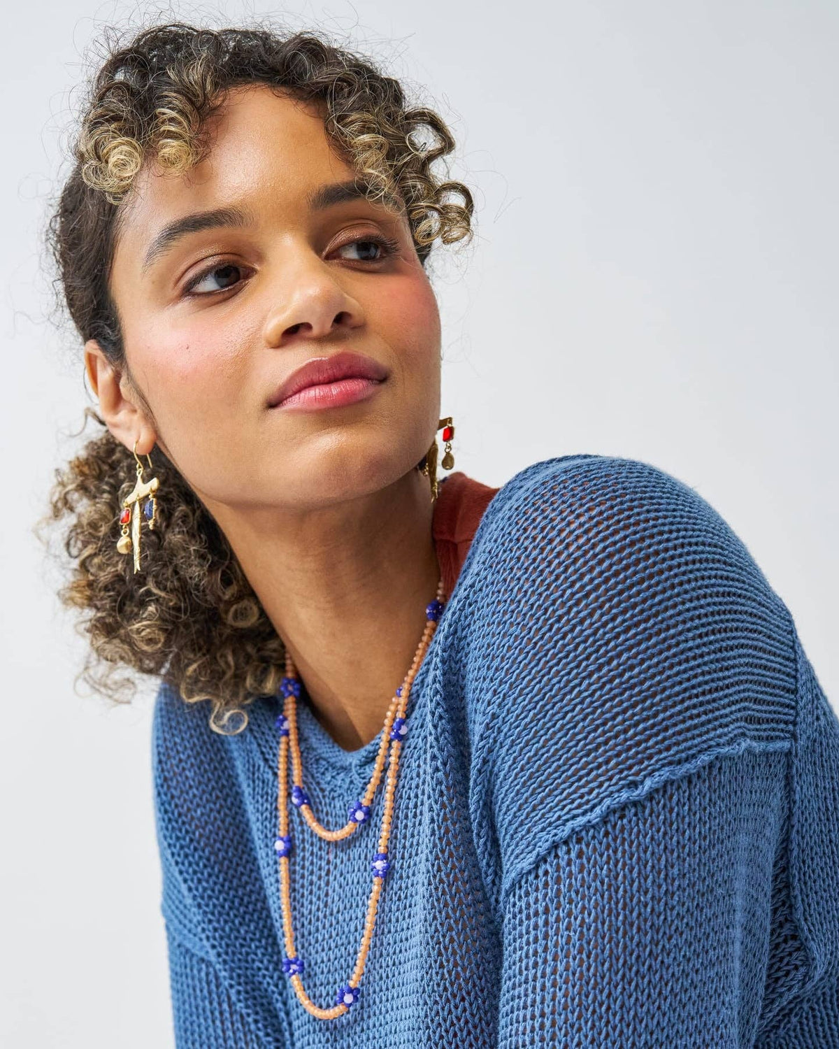 A woman with curly brown hair wearing a blue knit sweater, layered beaded necklaces, and colorful earrings, photographed against a light gray studio background.