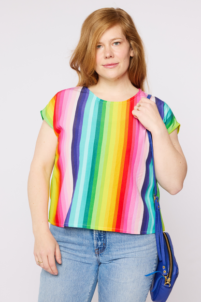 A person wearing a multicolored striped round neck cap sleeve top, paired with jeans, standing against a white background.