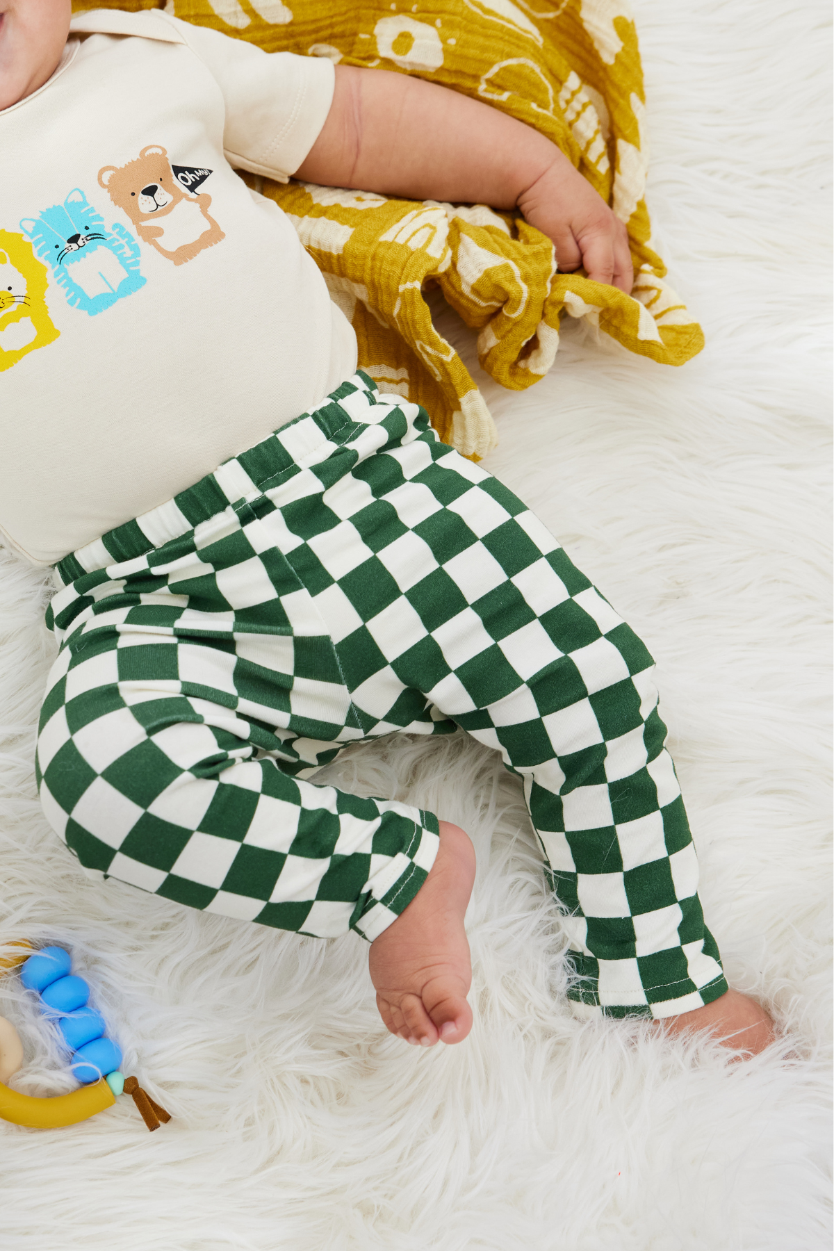 chubby 7 months old baby lying on a white furry rug wearing a onesie and green and white checker leggings.
